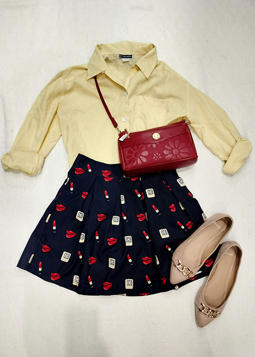 Flatlay of soft yellow shirt, patterned black skirt with deep red handbag and cream pumps