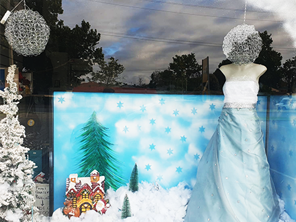Snow globe window display - fetauring a snow queen mannequin dressed in blue gown and icy cloak, there is a background painted with winter forests and a ginger bread house sitting on a pile of "snow" (made out of cushion stuffing).