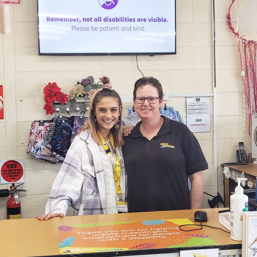 Two Good Sammy employees smiling at camera. The sign behind them says: Remember, not all disabilities are visible. Please be patient and kind.