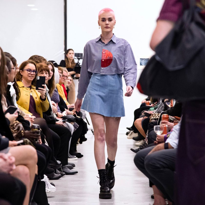 Person walking down runway with short pink hair wearing skirt, boots and jacket. People in front row watching, smiling and taking photos.