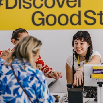 A staff member standing at the counter helping a customer. In the background is a yellow sign with black writing that says # Discover Good Stuff.