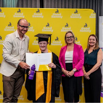Four people standing in a row and smiling at the camera. One person is wearing a black graduation gown and cap, and yellow sash, and is holding a folder with graduation papers.