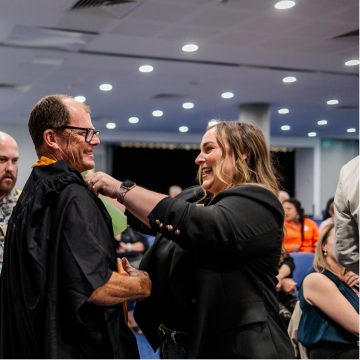 Two people facing each other and smiling. On the left is a man in a black graduation gown. On the right is a woman. She is adjusting the front of the gown.