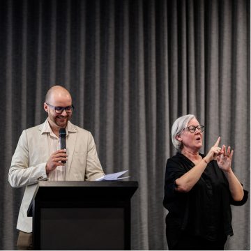 Two people addressing an audience. On the left, a man is holding a microphone and standing behind a lectern. On the right, a woman is signing using Auslan.