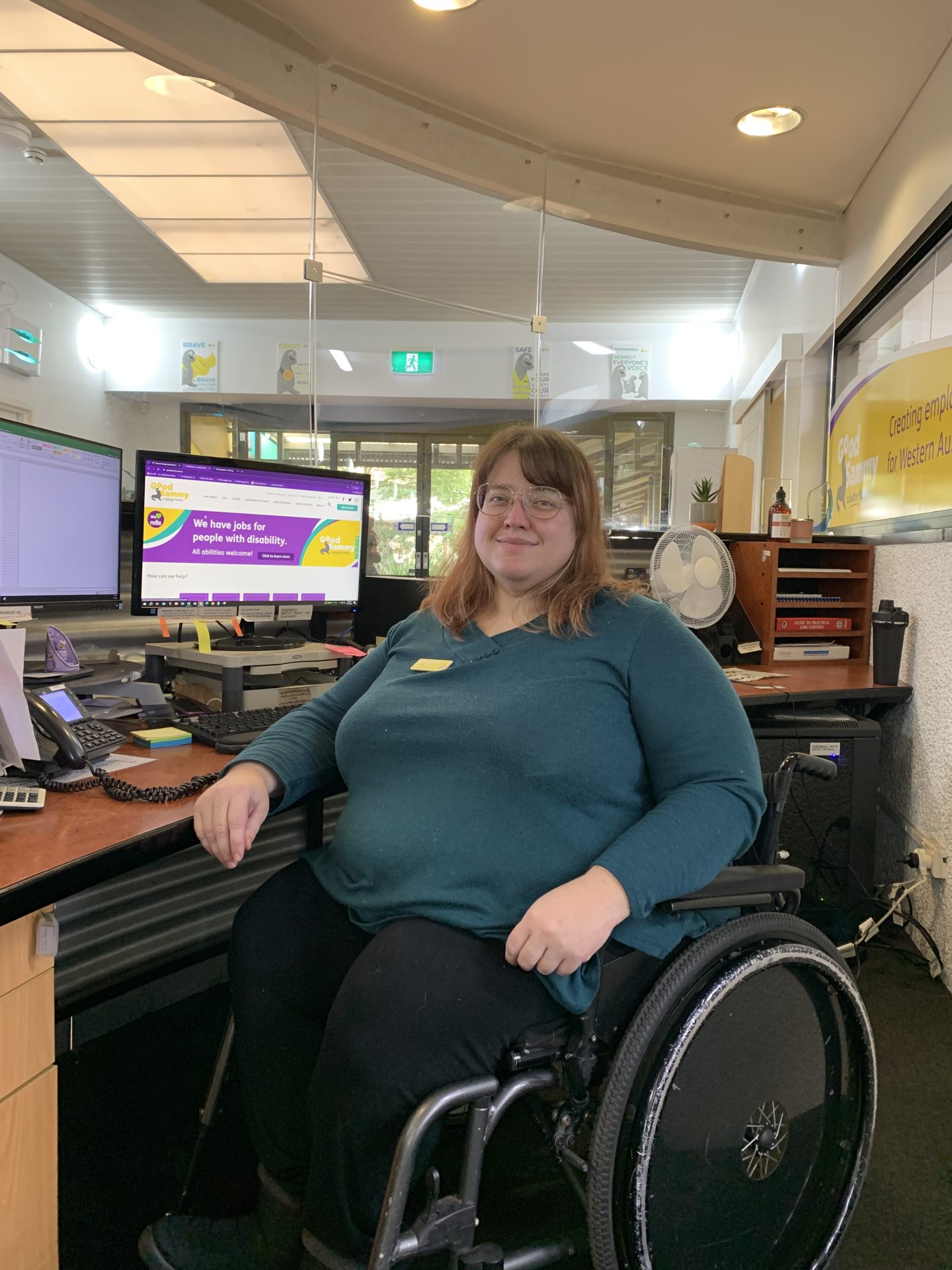 A person sitting at a desk in a wheelchair, smiling at the camera.